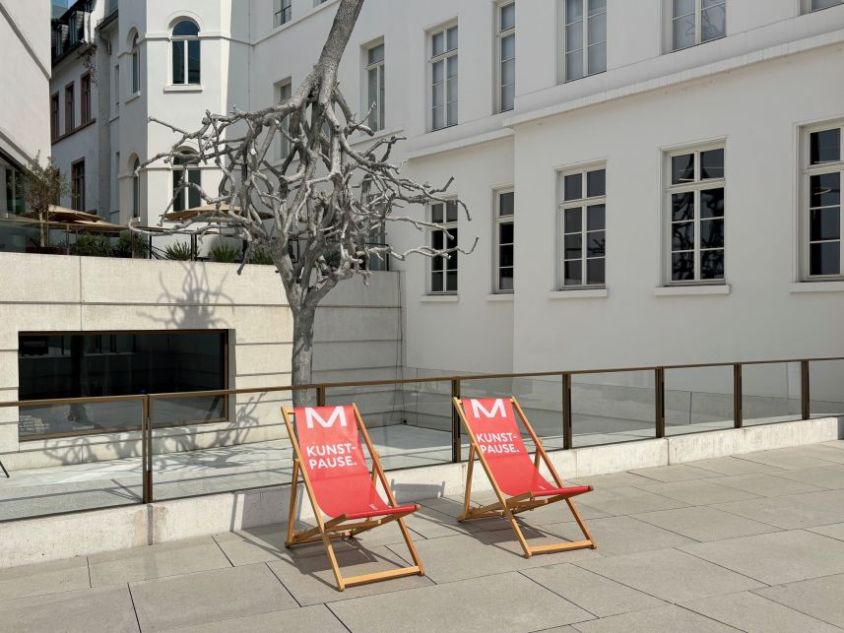 Zwei rote Liegest&uuml;hle mit der Aufschrift Kunstpause auf einem Platz vor einem Geb&auml;ude
