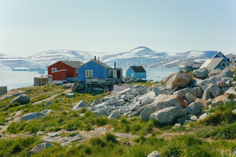 Kleine rote und blaue Holzh&auml;user an einer steinigen, grasbedeckten K&uuml;ste mit schneebedeckten H&uuml;geln im Hintergrund
