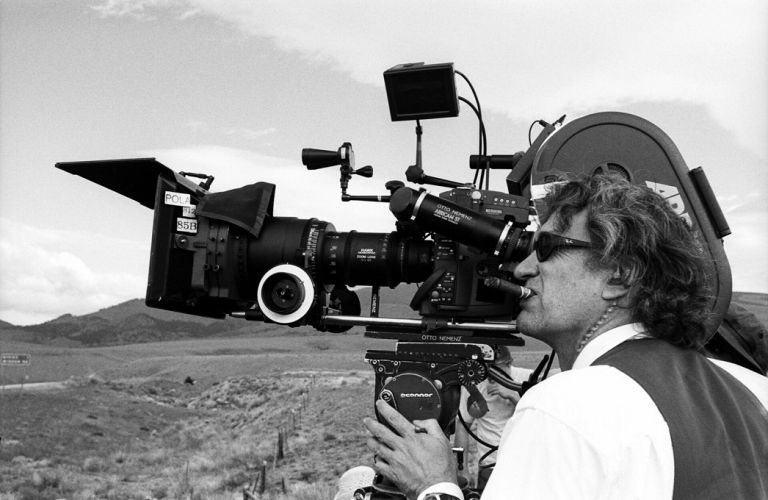 Schwarz-Wei&szlig;-Fotografie eines Mannes mit Sonnenbrille, der in einer kargen Landschaft steht und durch eine Filmkamera schaut